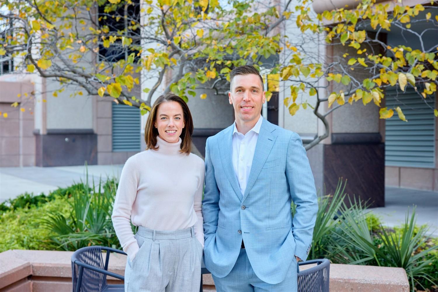 A professional outdoor portrait of a female executive in a white turtleneck and a male executive in a light blue suit standing in front of a WRMC corporate building.