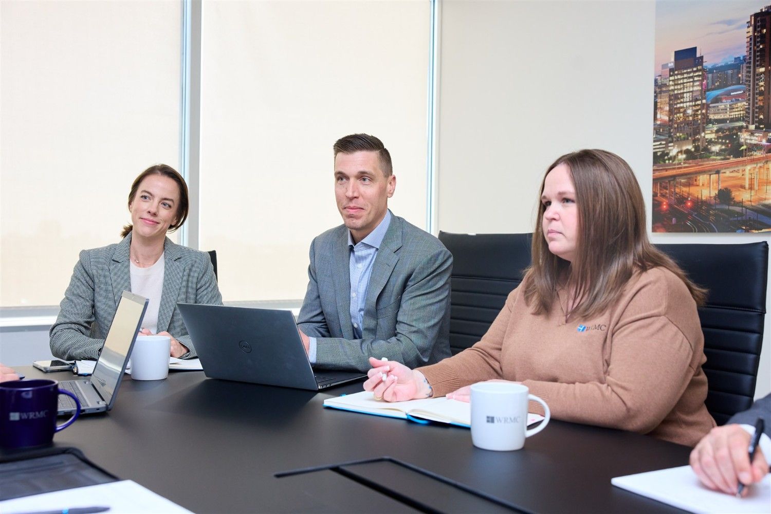 Three WRMC corporate executives sitting at a conference table with laptops and notebooks, listening attentively during a professional business presentation.