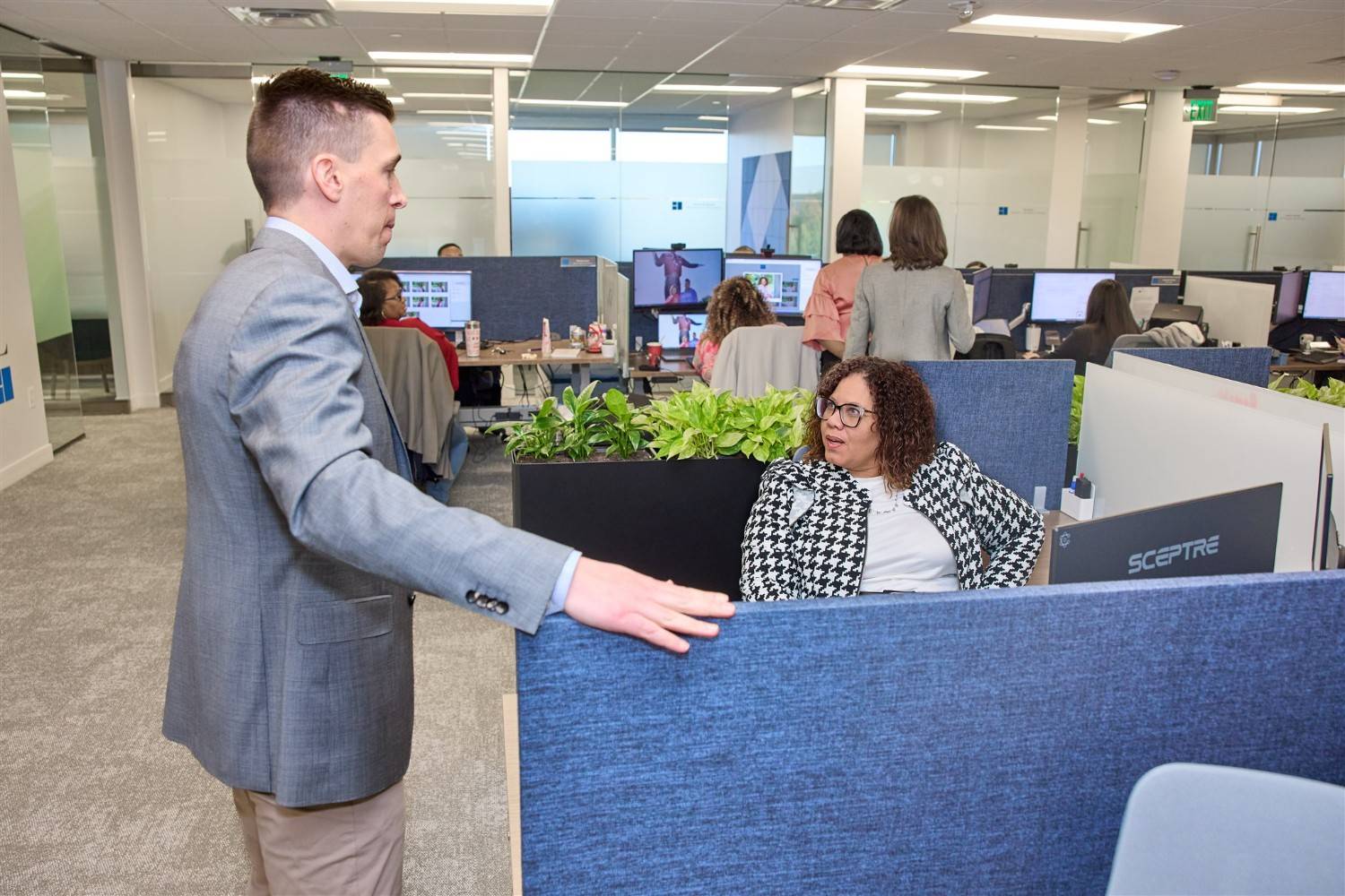 A male WRMC executive standing and speaking with a female colleague sitting at her cubicle desk in a busy open-concept corporate office.