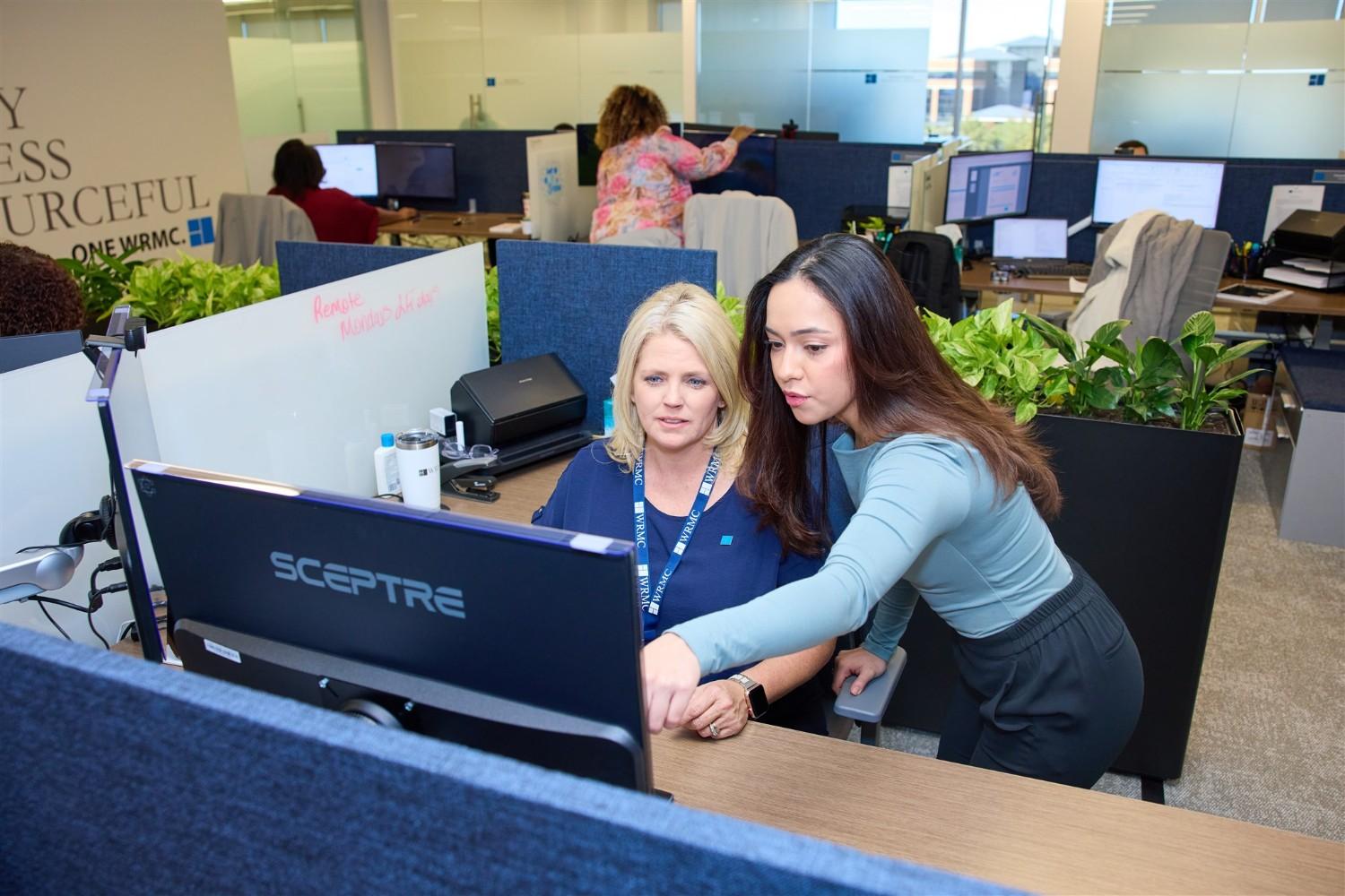 A female WRMC employee pointing to a Sceptre monitor to assist a coworker wearing a blue WRMC-branded lanyard at an office workstation.